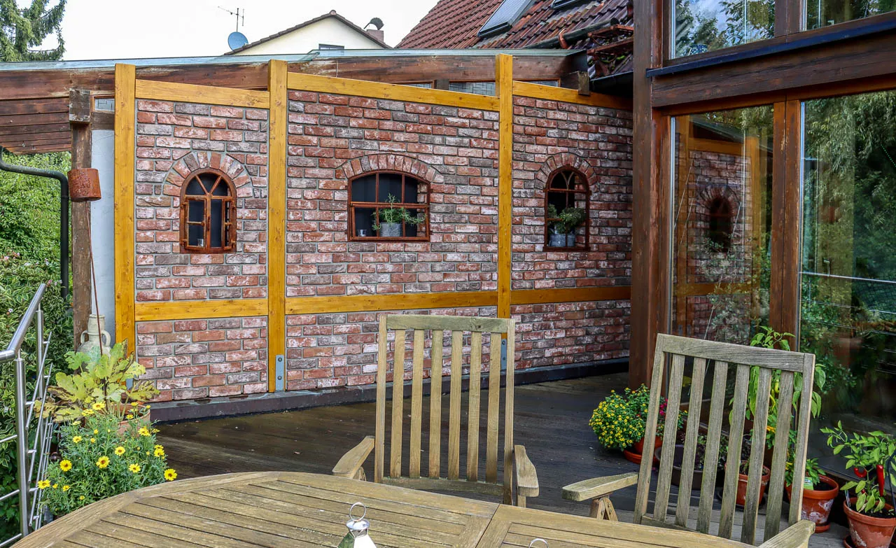 Sichtschutz-Backsteinwand auf der Terrasse. Die Wand mit rot braunen Klinkerriemchen passt super zu den Holzpfählen und den Gartenmöbeln aus Holz. 